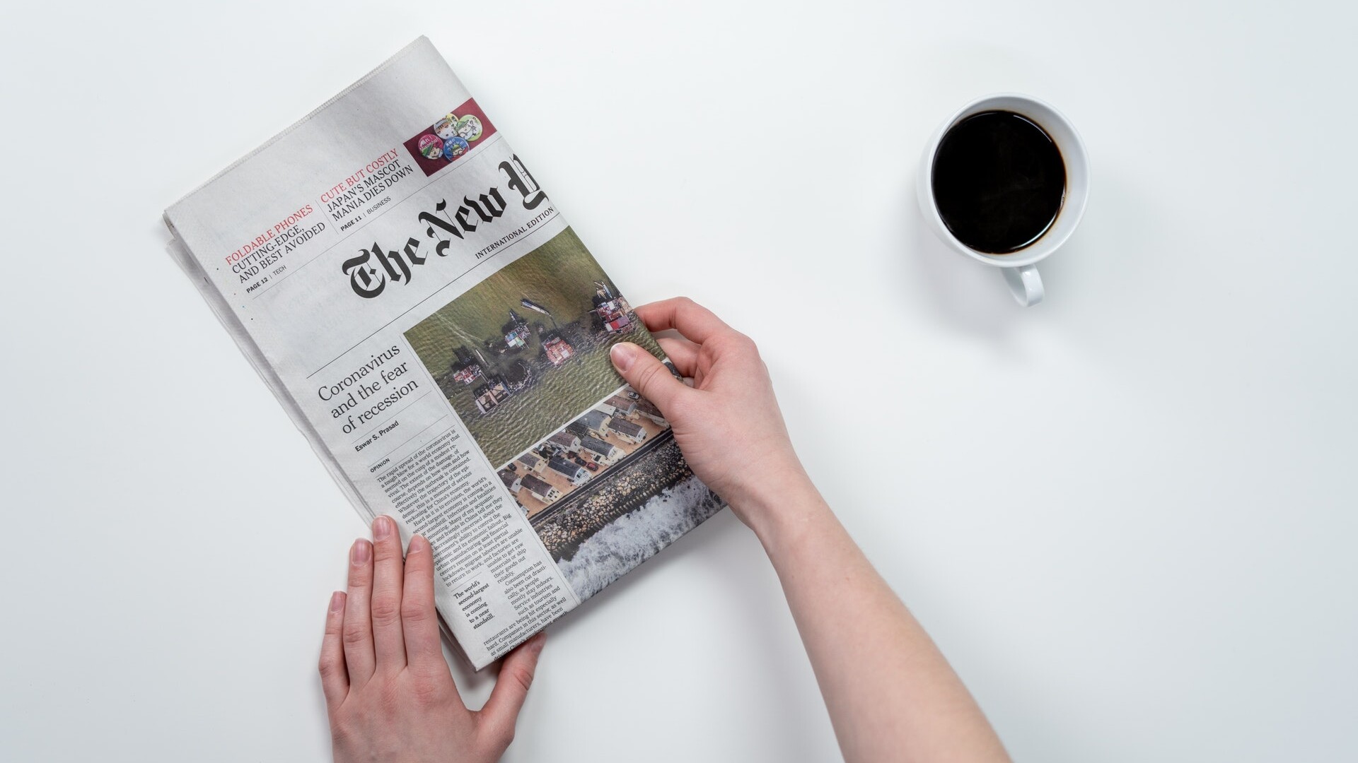 hands holding a folded newspaper with a cup coffee next to it